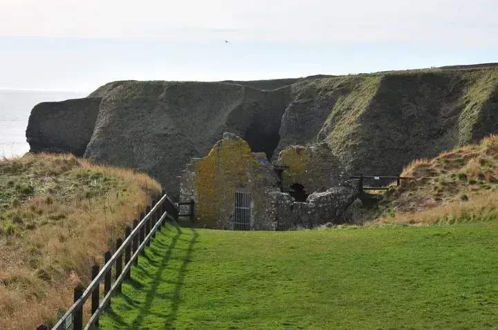 Low ruins and a fenced grassy path at Dunnottar Castle below coastal cliffs
