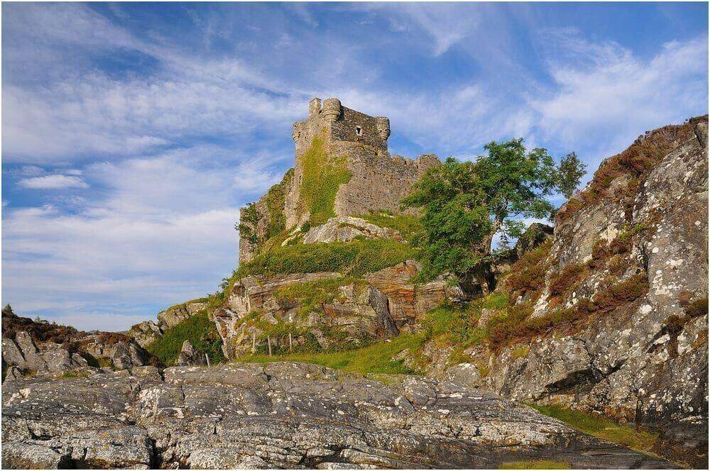 Tioram Castle ruins rising above layered rocks and shoreline trees